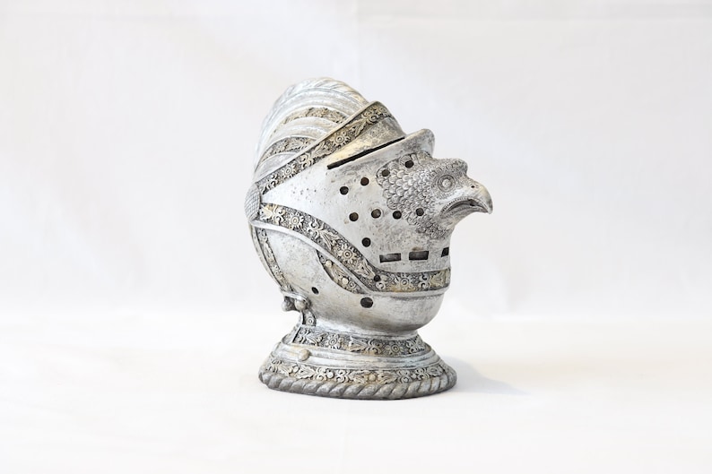 May include: A silver knight's helmet piggy bank with an eagle head design. The helmet has intricate details and a slot on the top for coins.