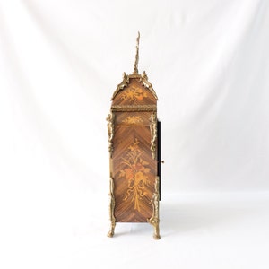 May include: A decorative clock with a marquetry design and gold accents. The clock has a floral motif and a black face. The clock is standing on four gold feet.