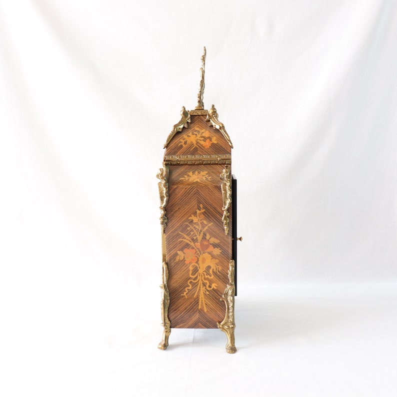 May include: A decorative clock with a marquetry design and gold accents. The clock has a floral motif and a black face. The clock is standing on four gold feet.