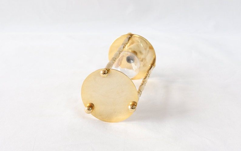Pu&ograve; includere: Una clessidra color oro con un centro in vetro trasparente. La clessidra ha una base e una parte superiore circolari, collegate da supporti curvi e ornati. La base ha tre piccoli piedini arrotondati. La clessidra &egrave; progettata per misurare il tempo.