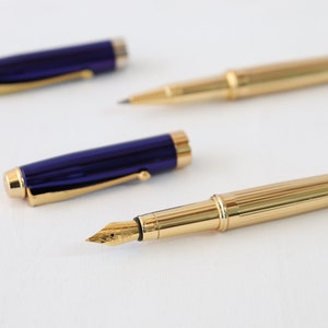 May include: Two fountain pens, one with a gold body and a gold nib, and the other with a blue body and a gold nib.