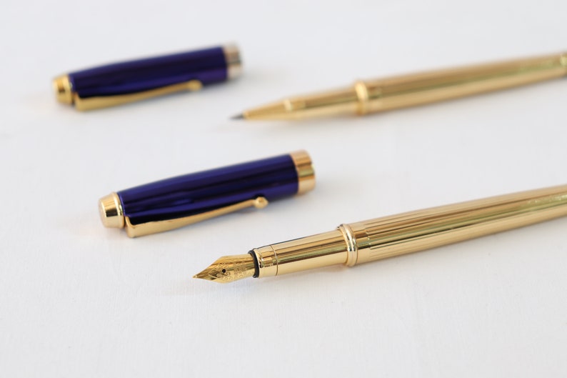 May include: Two fountain pens, one with a gold body and a gold nib, and the other with a blue body and a gold nib.