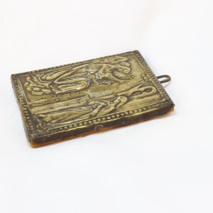 May include: A small, rectangular, brass icon with a raised design of two figures. The icon is mounted on a piece of brown fabric and has a small metal loop at the top.