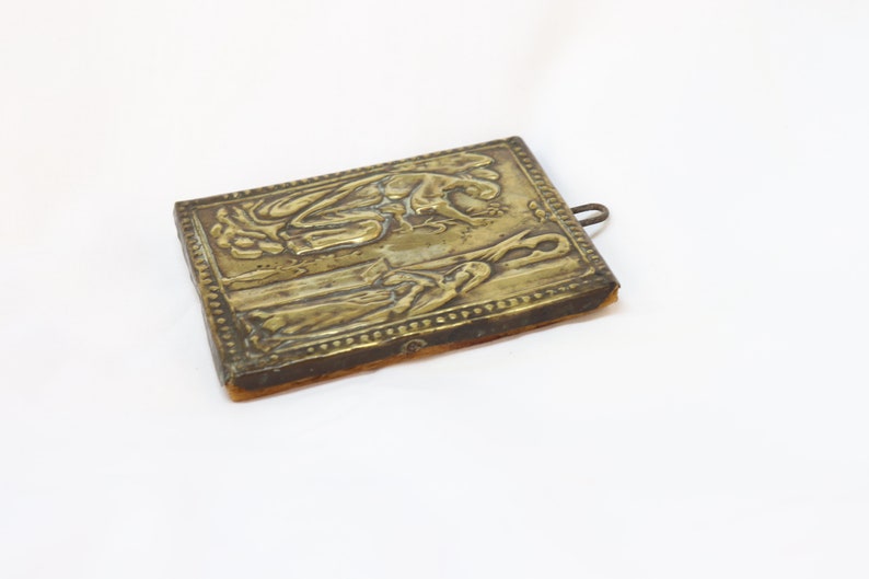 May include: A small, rectangular, brass icon with a raised design of two figures. The icon is mounted on a piece of brown fabric and has a small metal loop at the top.
