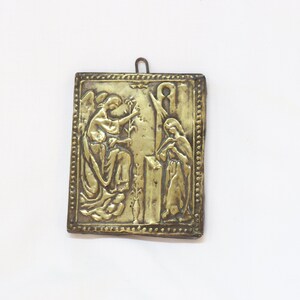 May include: A gold-colored metal plaque with an embossed image of an angel and a woman. The plaque has a decorative border and a loop at the top for hanging.