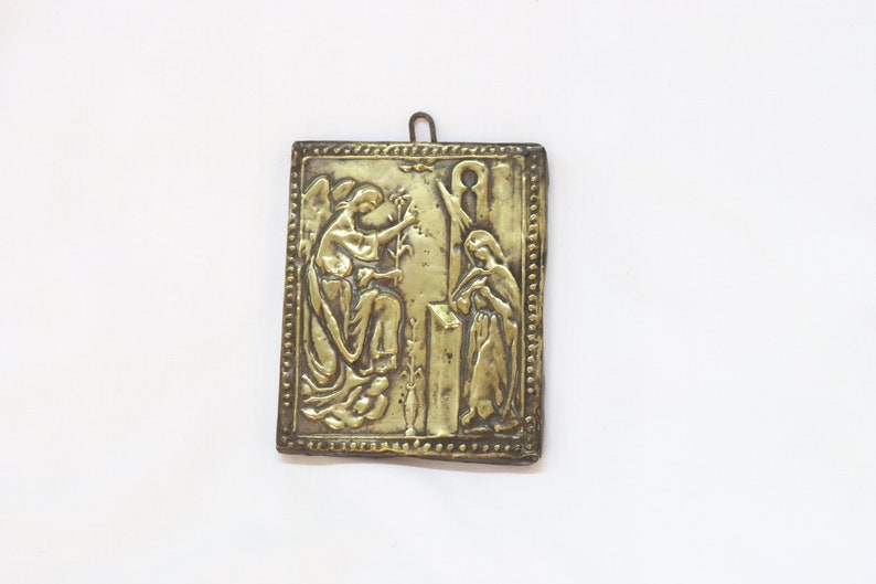 May include: A gold-colored metal plaque with an embossed image of an angel and a woman. The plaque has a decorative border and a loop at the top for hanging.