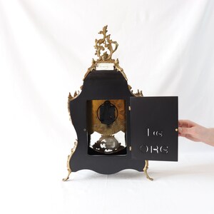 May include: A gold-toned ornate clock with a black face and a small door on the side. The clock has a decorative design with intricate details.