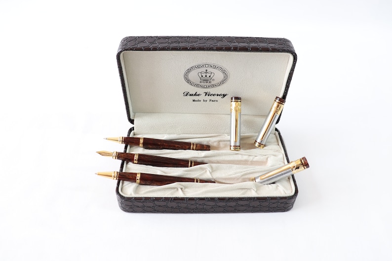 May include: A set of five pens in a brown crocodile-embossed box. The pens are made of wood and metal with gold accents. The box has a label that reads "Duke Viceroy Made by Paris."
