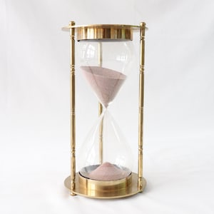 May include: A brass hourglass with a glass bulb filled with pink sand. The hourglass is on a white surface.