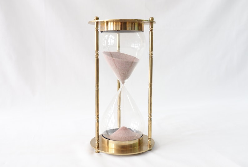May include: A brass hourglass with a glass bulb filled with pink sand. The hourglass is on a white surface.