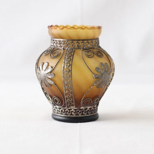 May include: A small amber glass vase with a decorative metal design featuring floral patterns. The vase has a scalloped rim and a black base.