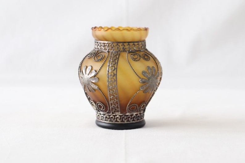 May include: A small amber glass vase with a decorative metal design featuring floral patterns. The vase has a scalloped rim and a black base.