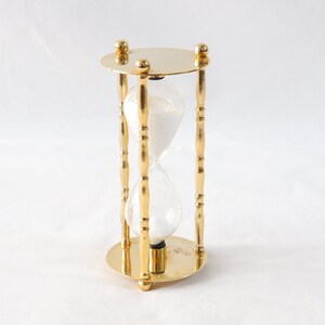 Pu&ograve; includere: Una clessidra color oro con un centro in vetro trasparente riempito di sabbia bianca. La clessidra ha una base e una parte superiore circolari, collegate da quattro supporti verticali decorati. Il design &egrave; simmetrico.