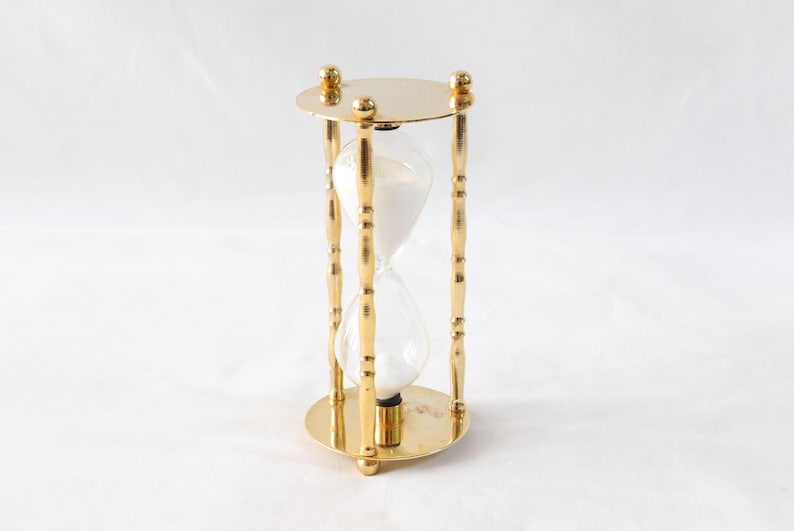 Pu&ograve; includere: Una clessidra color oro con un centro in vetro trasparente riempito di sabbia bianca. La clessidra ha una base e una parte superiore circolari, collegate da quattro supporti verticali decorati. Il design &egrave; simmetrico.