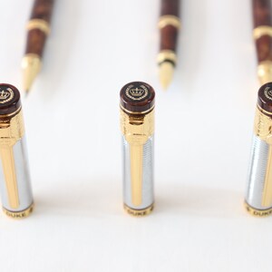 May include: Three silver and gold pens with brown wood accents and a crown logo on the top. The pens are on a white surface.