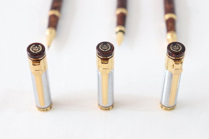 May include: Three silver and gold pens with brown wood accents and a crown logo on the top. The pens are on a white surface.