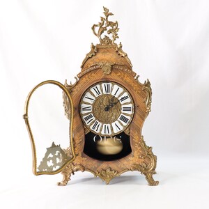 May include: A vintage, ornate, wooden mantel clock with a gold-toned finish and a glass door. The clock face has Roman numerals and a pendulum.