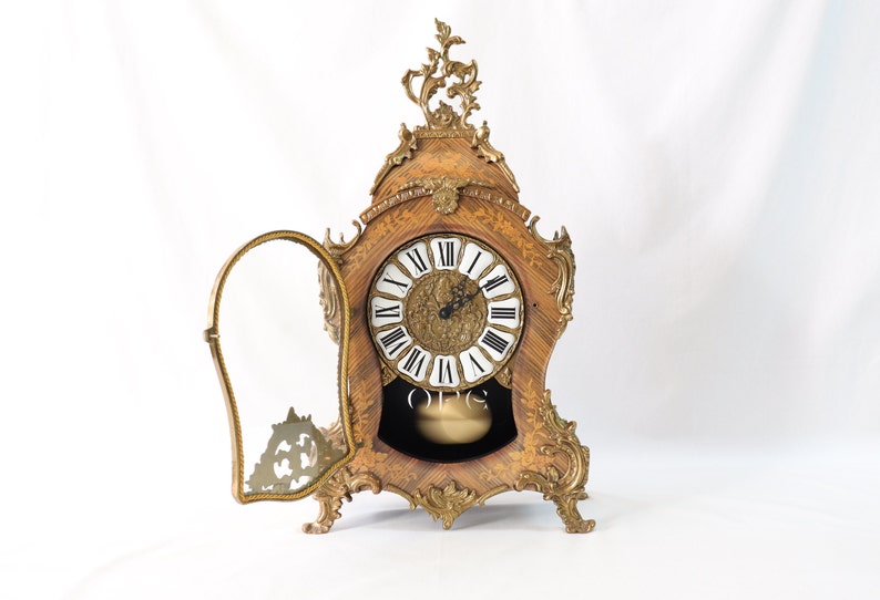 May include: A vintage, ornate, wooden mantel clock with a gold-toned finish and a glass door. The clock face has Roman numerals and a pendulum.