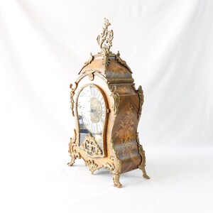 May include: A vintage, ornate, wooden mantel clock with a gold-toned finish and intricate carvings. The clock face is white with black Roman numerals and a gold-toned hour and minute hand.