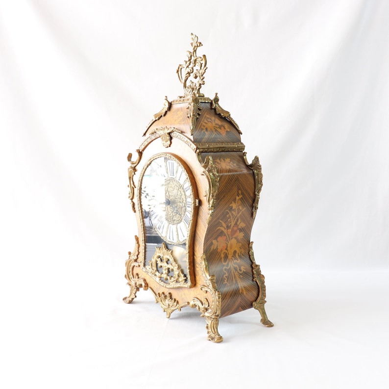 May include: A vintage, ornate, wooden mantel clock with a gold-toned finish and intricate carvings. The clock face is white with black Roman numerals and a gold-toned hour and minute hand.