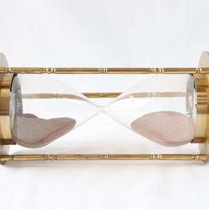 May include: A brass hourglass with a clear glass bulb and pink sand. The hourglass is on a white background.
