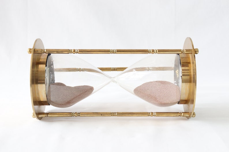 May include: A brass hourglass with a clear glass bulb and pink sand. The hourglass is on a white background.