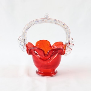 Può includere: Un cestino di vetro rosso con un manico di vetro trasparente. Il cestino ha un bordo arricciato ed è decorato con macchie bianche e blu.