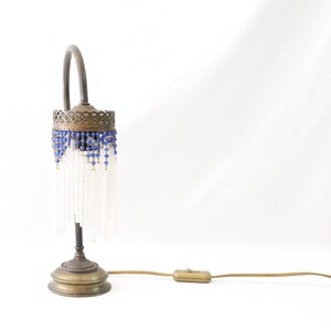 K&ouml;nnte beinhalten: Eine Tischlampe aus Messing mit einem wei&szlig;en Perlenbesatz und blauen Glasperlen. Die Lampe hat einen gebogenen Arm und einen runden Sockel.