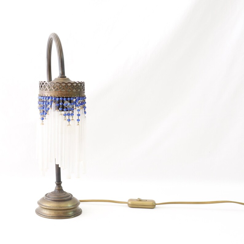 K&ouml;nnte beinhalten: Eine Tischlampe aus Messing mit einem wei&szlig;en Perlenbesatz und blauen Glasperlen. Die Lampe hat einen gebogenen Arm und einen runden Sockel.