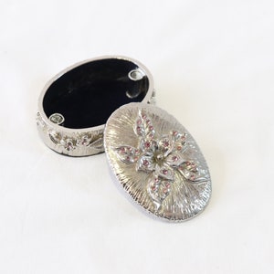 May include: A silver oval trinket box with a floral design. The lid features a raised flower with pink rhinestones. The box is open and the inside is lined with black velvet.