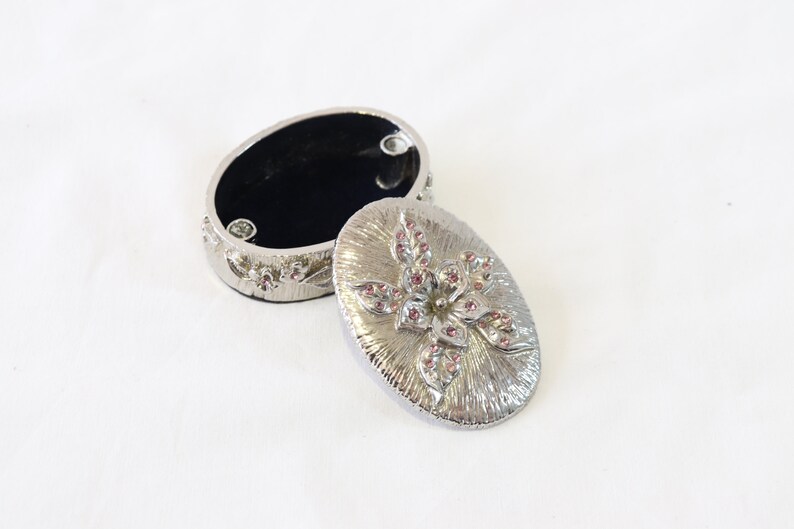 May include: A silver oval trinket box with a floral design. The lid features a raised flower with pink rhinestones. The box is open and the inside is lined with black velvet.