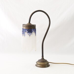 K&ouml;nnte beinhalten: Eine Tischlampe aus Messing mit einem wei&szlig;en Perlenlampenschirm und blauen Akzenten. Die Lampe hat einen gebogenen Arm und einen runden Sockel.