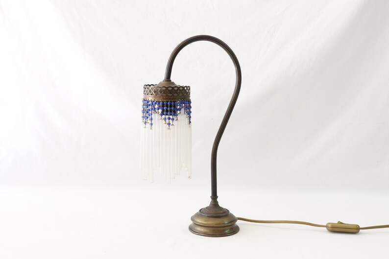 K&ouml;nnte beinhalten: Eine Tischlampe aus Messing mit einem wei&szlig;en Perlenlampenschirm und blauen Akzenten. Die Lampe hat einen gebogenen Arm und einen runden Sockel.