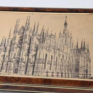 May include: A framed black and white line drawing of the Duomo di Milano, the cathedral in Milan, Italy. The drawing is on a beige background and is framed in a dark wood frame with a gold trim.