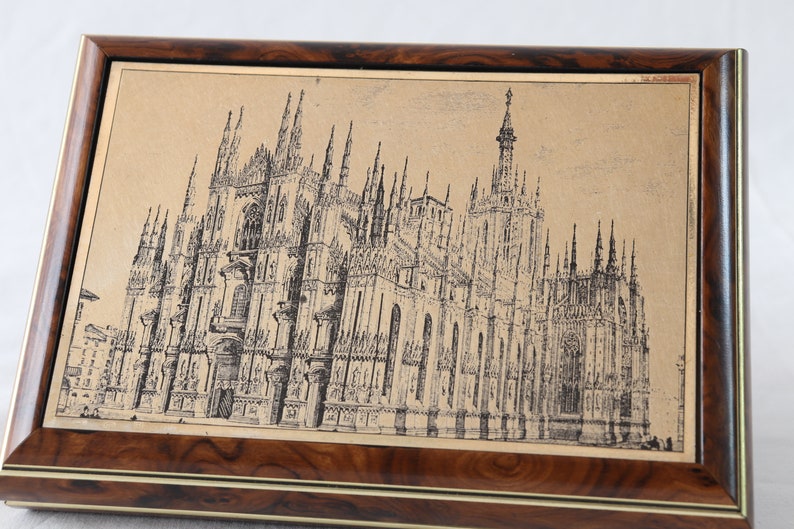 May include: A framed black and white line drawing of the Duomo di Milano, the cathedral in Milan, Italy. The drawing is on a beige background and is framed in a dark wood frame with a gold trim.