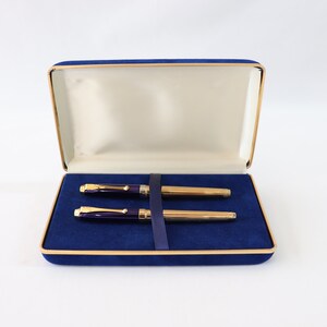May include: A blue velvet pen case with a gold trim, containing two gold and purple pens.