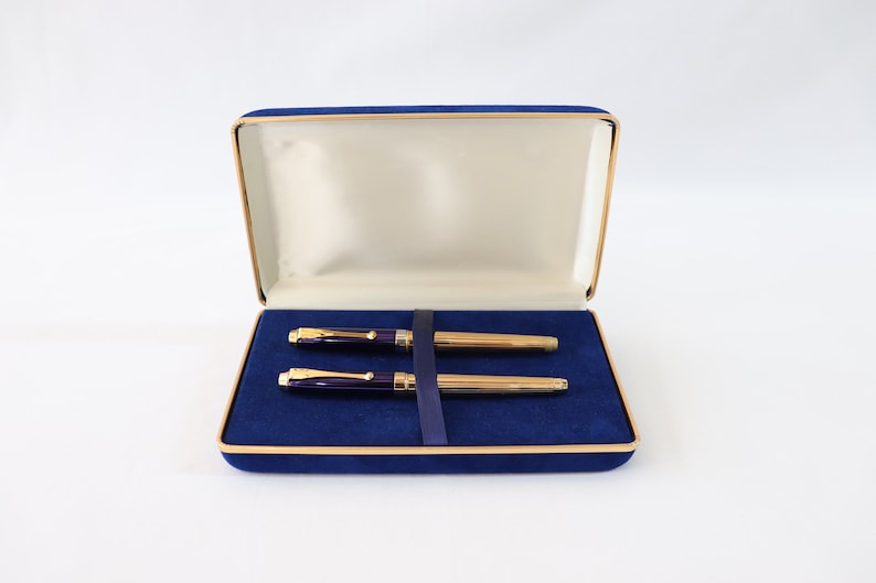 May include: A blue velvet pen case with a gold trim, containing two gold and purple pens.