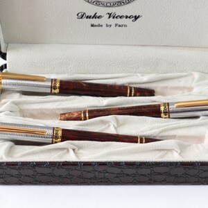May include: A set of three brown and silver pens with gold accents in a brown leather case. The pens are labeled "Duke Viceroy" and "Made by Farn".
