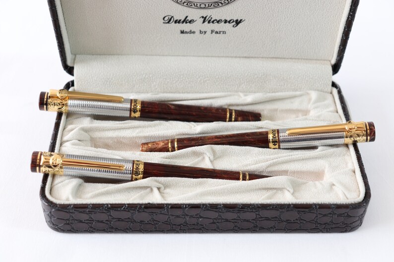 May include: A set of three brown and silver pens with gold accents in a brown leather case. The pens are labeled "Duke Viceroy" and "Made by Farn".