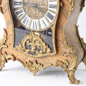 May include: A decorative, ornate, gold-toned clock with a black face and Roman numeral markings. The clock has intricate carvings and a wood grain finish.