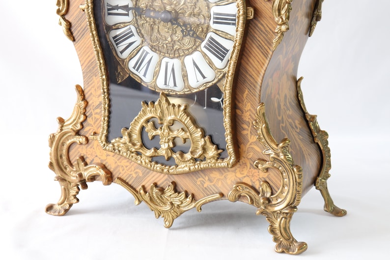 May include: A decorative, ornate, gold-toned clock with a black face and Roman numeral markings. The clock has intricate carvings and a wood grain finish.