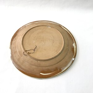 May include: A brown ceramic plate with a white rim and a brown glaze. The plate has a hole in the top for hanging. The back of the plate has the word "KOROND" written on it.