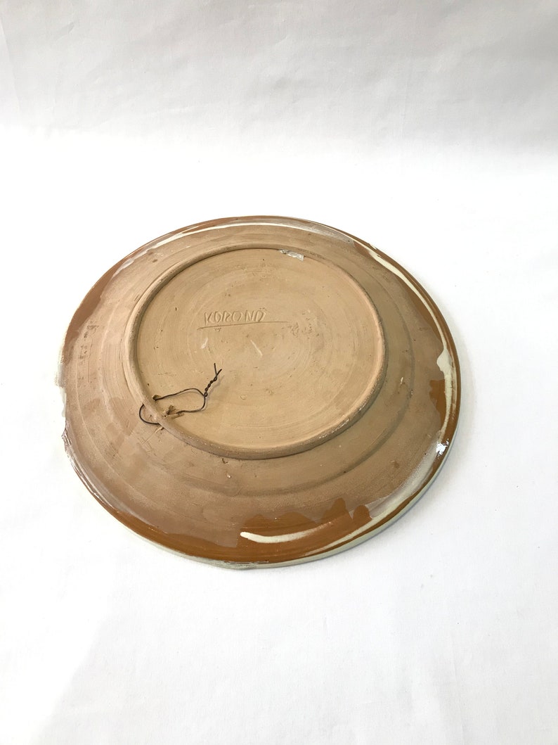 May include: A brown ceramic plate with a white rim and a brown glaze. The plate has a hole in the top for hanging. The back of the plate has the word "KOROND" written on it.