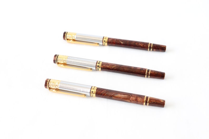 May include: Three brown and silver pens with gold accents. The pens have a wood-like finish and are arranged in a row.