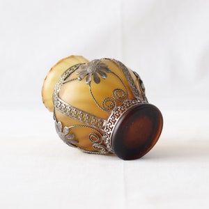 May include: A small, amber-colored glass vase with a silver metal filigree design. The vase has a round body and a wide mouth.