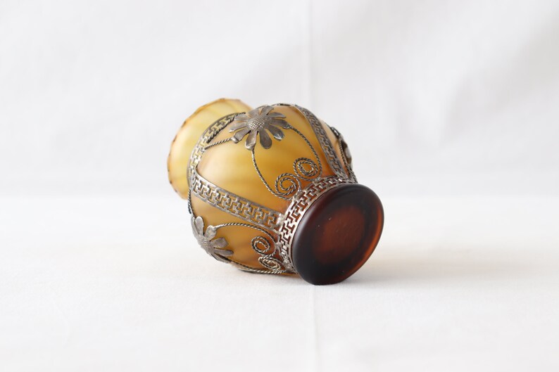 May include: A small, amber-colored glass vase with a silver metal filigree design. The vase has a round body and a wide mouth.