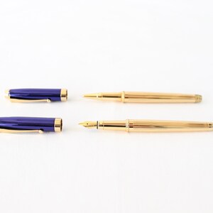 May include: A set of two pens, one with a blue barrel and a gold cap and the other with a gold barrel and a gold cap.