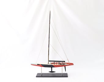 May include: A red, black and white model sailboat with a black mast and white sails. The model is on a black stand.