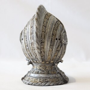 May include: A silver and gold helmet with intricate details and a pointed top. The helmet is made of metal and has a textured surface.