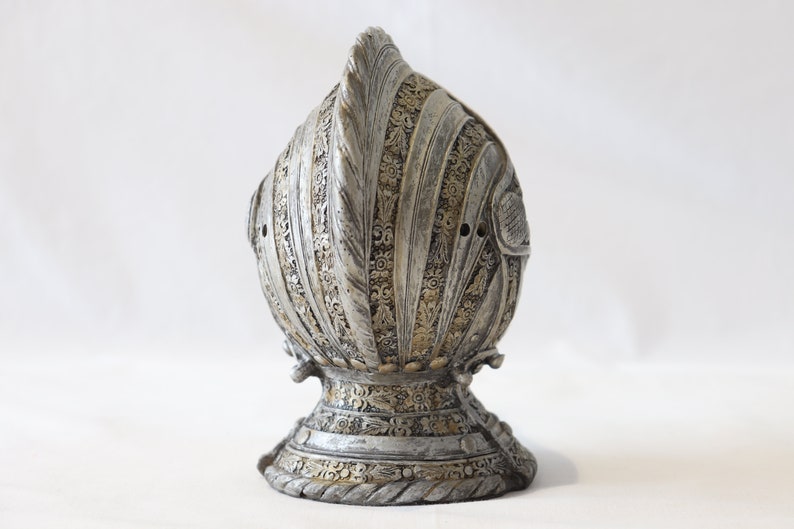 May include: A silver and gold helmet with intricate details and a pointed top. The helmet is made of metal and has a textured surface.